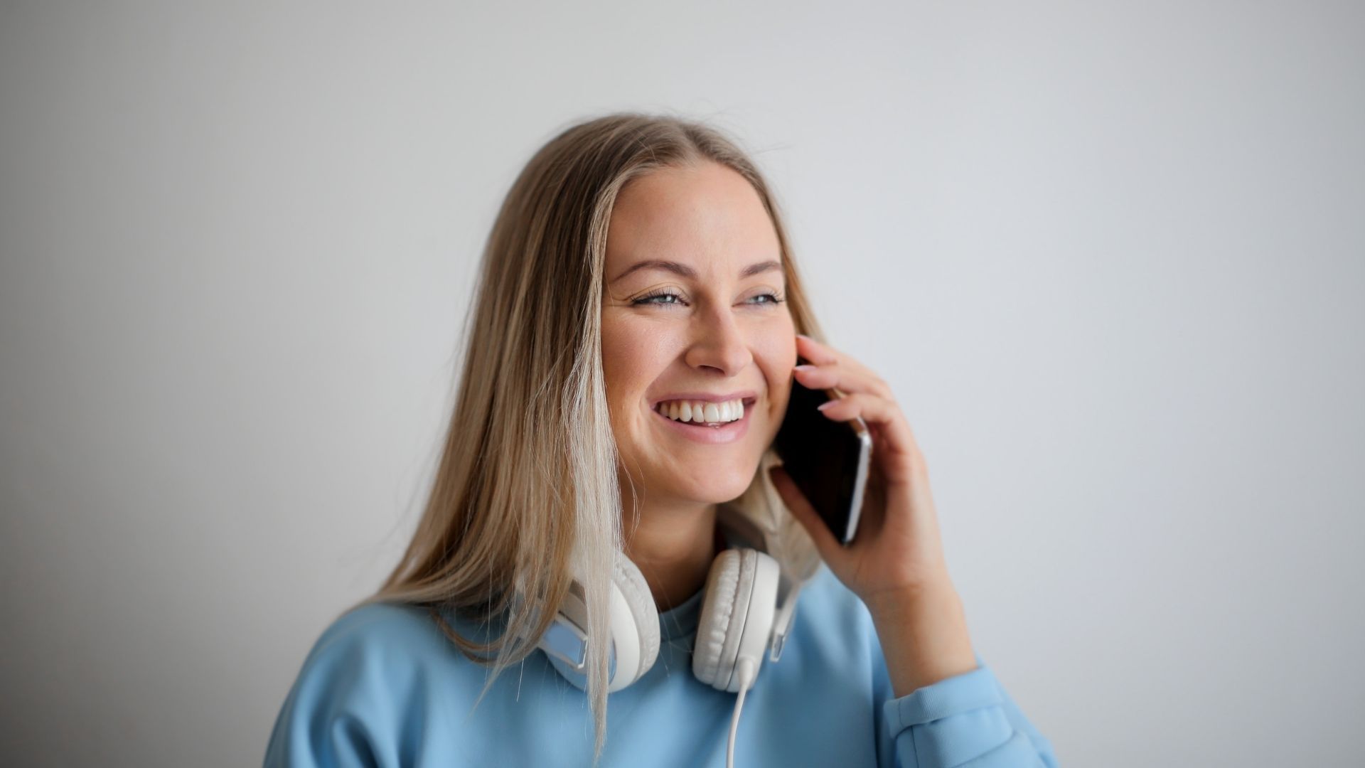 Young woman talking on phone wearing headphones, blue hoodie, neutral background. potential pitfalls in digital communication, network congestion